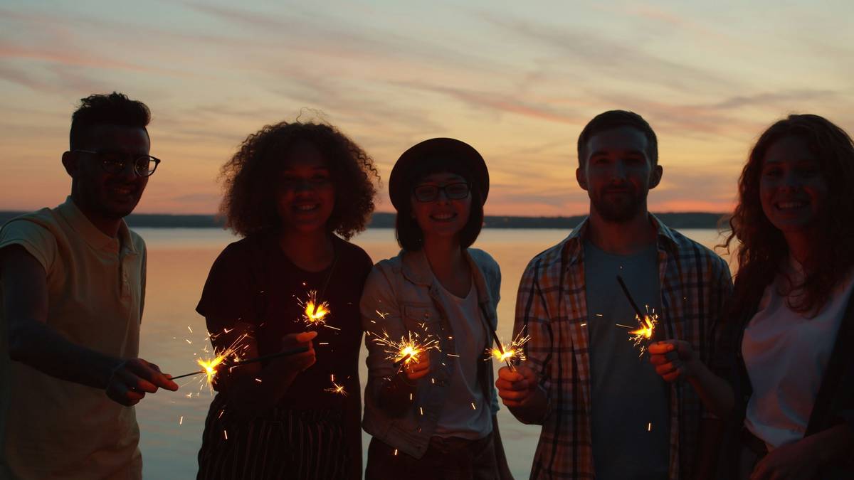 Portrait of happy young people holding bengal lights standing on beach in the evening together smiling looking at camera. Lifestyle and festivals concept.